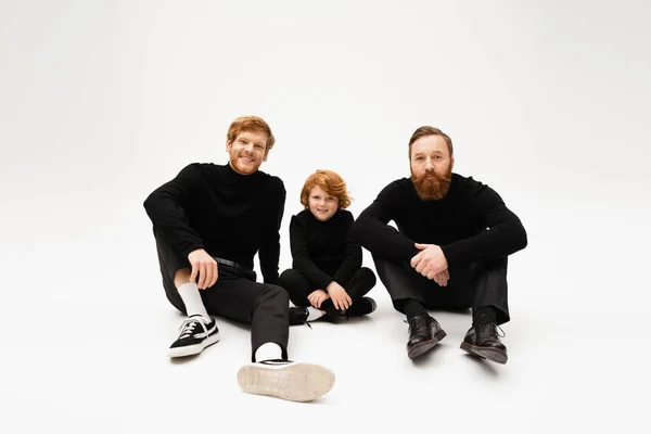 Full length of redhead men in black clothes posing near cheerful boy sitting with crossed legs on light grey background — Stock Photo