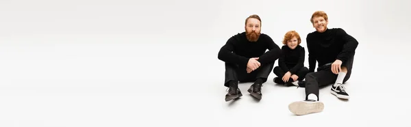 Full length of adult bearded men and red haired kid sitting in black clothes on light grey background, banner — Stock Photo