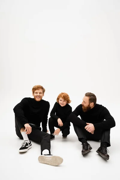 Cheerful man smiling at camera while sitting near redhead son and bearded dad on light grey background — Photo de stock