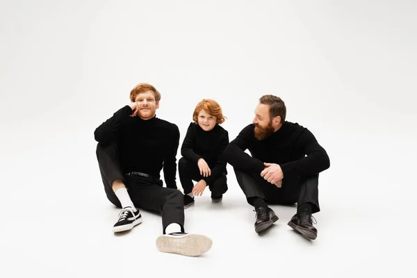 Joyful redhead man sitting near smiling son and bearded father in black clothes on light grey background — Stock Photo
