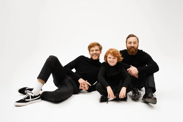 Happy bearded men in black clothes smiling at camera near redhead boy while posing on light grey background — Stock Photo