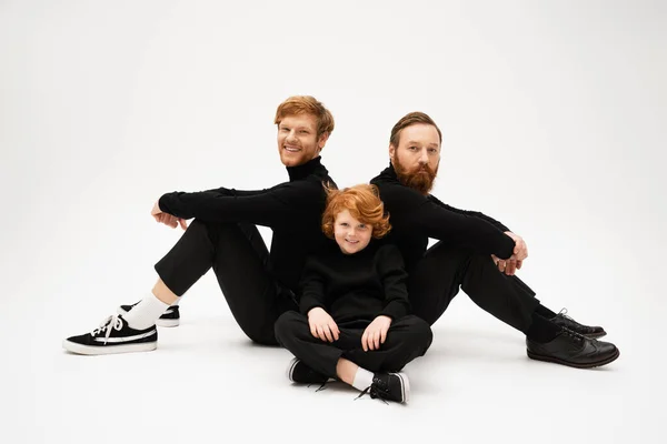 Smiling bearded men in black turtlenecks sitting back to back near happy red haired kid on light grey background — Stock Photo