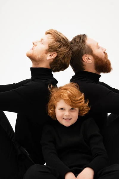 Red haired boy smiling at camera near bearded grandpa and father in black turtlenecks sitting back to back isolated on grey — Stock Photo