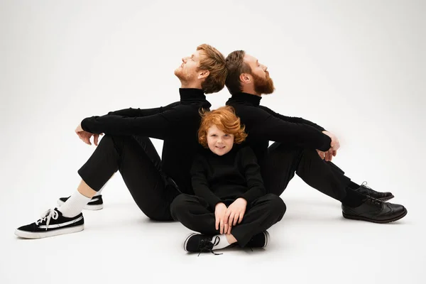 Redhead boy smiling at camera near bearded grandfather and dad sitting back to back on light grey background — Foto stock