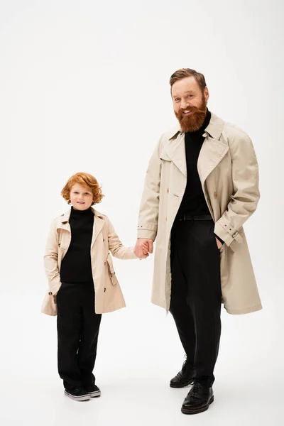 Full length of cheerful red haired boy with grandfather holding hands while posing in trench coats on light grey background — Foto stock