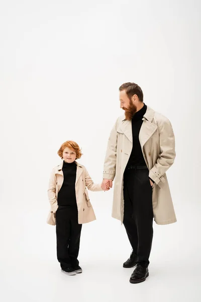 Full length of bearded man in trench coat holding hands with redhead grandson posing with hand in pocket on light grey background — Foto stock