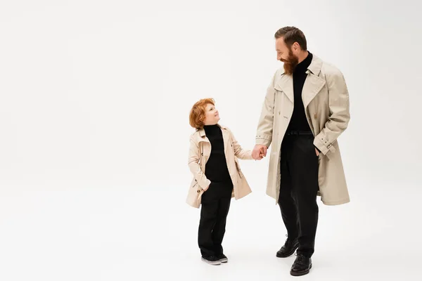 Happy redhead kid and bearded man in trench coats holding hands and looking at each other on light grey background — Stock Photo