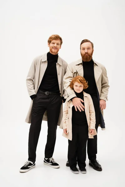 Full length of smiling red haired boy with father and grandpa in trench coats looking at camera on light grey background — Foto stock