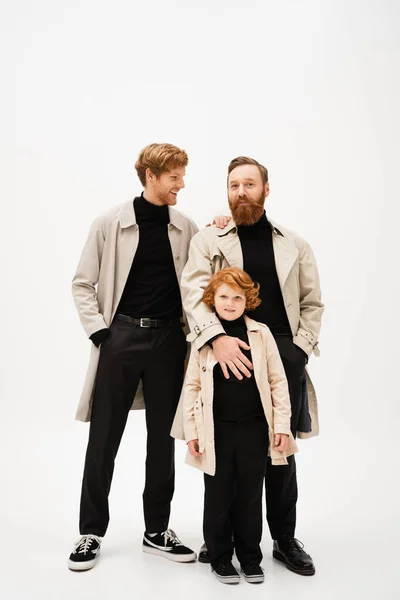Smiling man standing with hand in pocket near bearded dad and redhead son on light grey background — Stock Photo