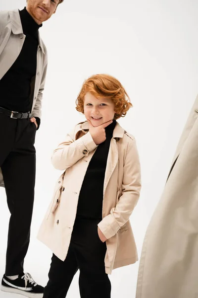 Happy redhead boy in trench coat touching chin while posing with hand in pocket on light grey background — Stock Photo