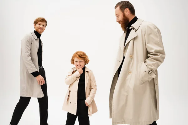 Cheerful redhead boy touching chin while posing with hand in pocket near dad and grandpa isolated on grey — Stock Photo