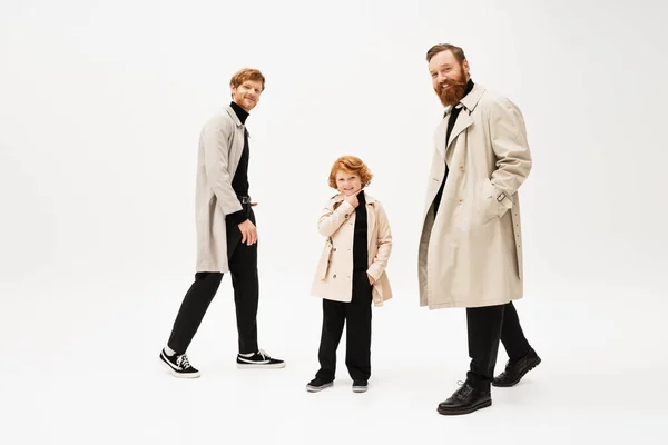 Full length of bearded man in trench coat smiling at camera near redhead son and grandson on light grey background — Stockfoto
