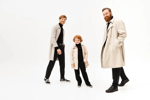 Full length of happy redhead men and kid in trench coats looking at camera while posing on light grey background — Stock Photo