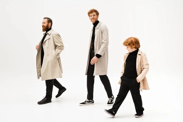 Young man in trench coat smiling at camera while walking near bearded father and redhead son on light grey background — Stock Photo