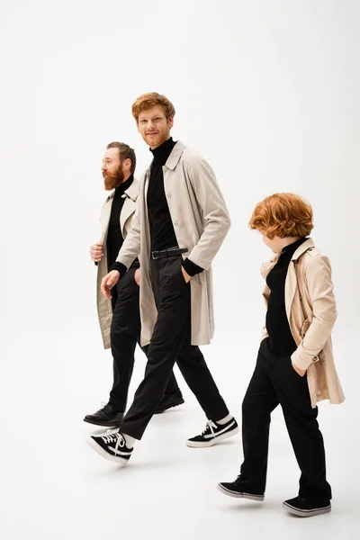 Full length of trendy bearded men and redhead boy in trench coats walking on light grey background — Foto stock
