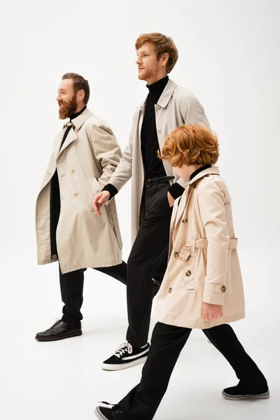 Bearded men and redhead boy in trench coats walking on light grey background — Foto stock