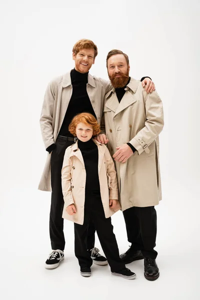 Cheerful man hugging bearded dad near redhead son while posing in trench coats on light grey background — Stock Photo