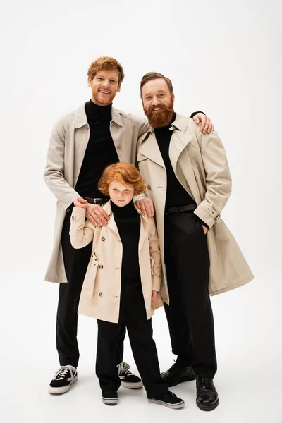 Joyful red haired man embracing bearded father and son standing in trench coats on light grey background — Stock Photo