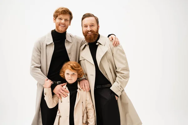 Happy bearded men and red haired kid in trench coats looking at camera isolated on grey — Stock Photo