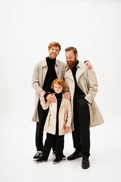 Happy redhead man embracing son and bearded father posing in trench coats on light grey background — Foto stock
