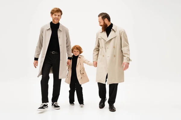 Full length of redhead men in trench coats holding hands with smiling boy while walking on light grey background — Photo de stock