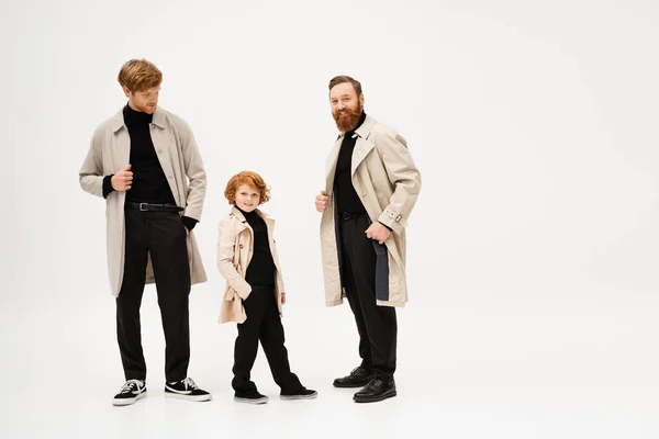 Red haired boy smiling at camera while standing with hand in pocket near dad and bearded grandpa in trench coats on light grey background — Foto stock