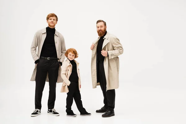 Smiling boy posing with hand in pocket near redhead dad and grandfather in trench coats on light grey background — Foto stock