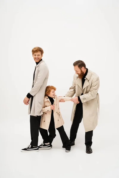 Full length of redhead man in trench coat smiling near bearded grandpa and grandson having fun on light grey background — Foto stock