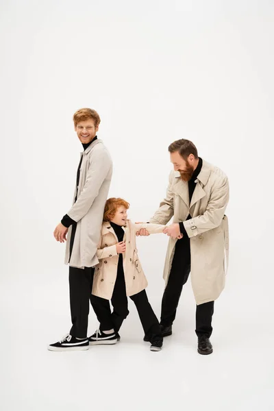 Full length of redhead man in trench coat smiling near bearded grandpa and grandson having fun on light grey background — Stock Photo