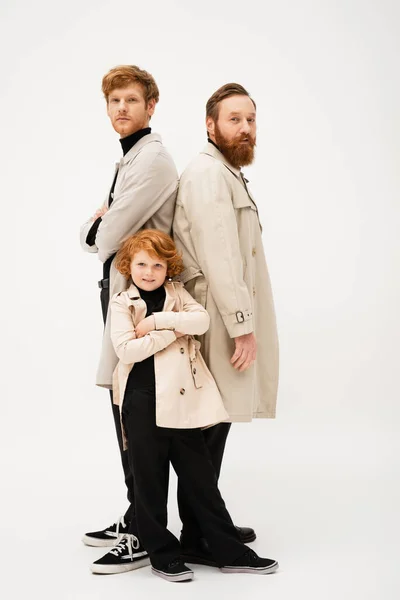 Happy red haired boy posing with crossed arms near father and grandpa in trench coats standing back to back on light grey background — Foto stock