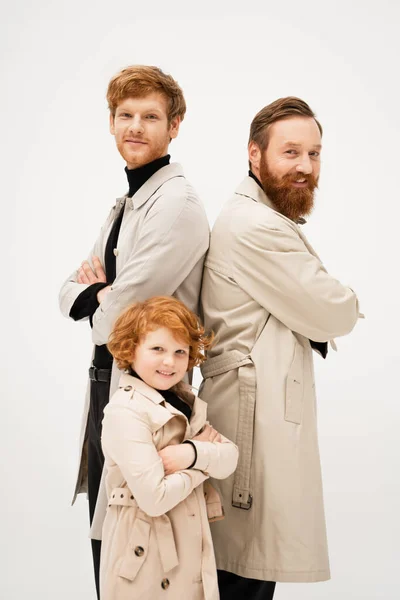 Cheerful redhead boy with father and grandpa in trench coats posing with crossed arms isolated on grey — Stock Photo