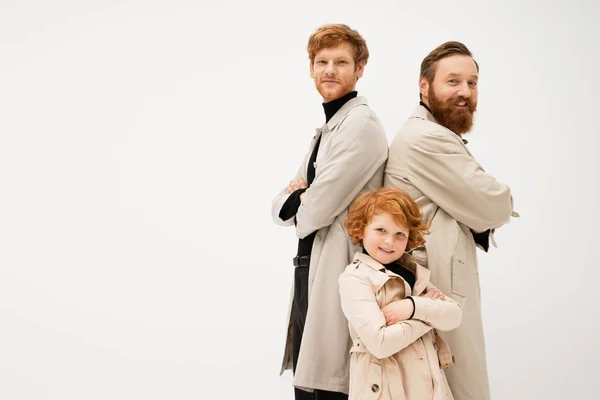 Joyful bearded man in trench coats standing back to back with crossed arms near redhead boy isolated on grey — Foto stock