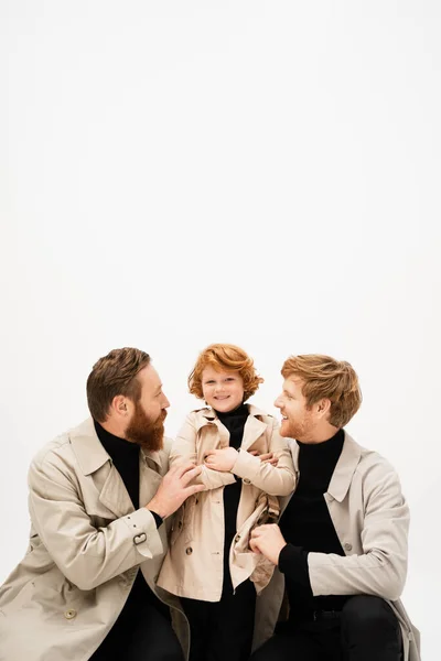Happy redhead kid in trench coat smiling near father and bearded grandpa hugging him isolated on grey — Stock Photo