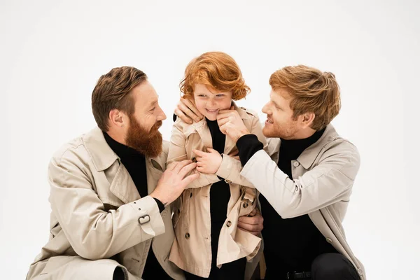 Happy bearded men in trench coats embracing cheerful redhead boy isolated on grey — Stock Photo