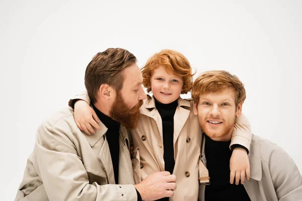 Smiling kid in trench coat hugging redhead father and bearded granddad isolated on grey — Stock Photo