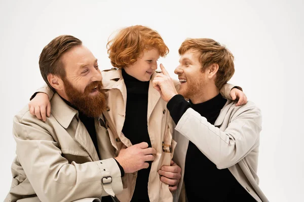 Cheerful redhead men in trench coats embracing and having fun with smiling boy isolated on grey — Stock Photo