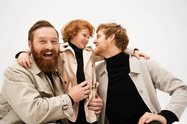 Joyful bearded man laughing at camera near redhead son and grandson in trench coats isolated on grey — Foto stock