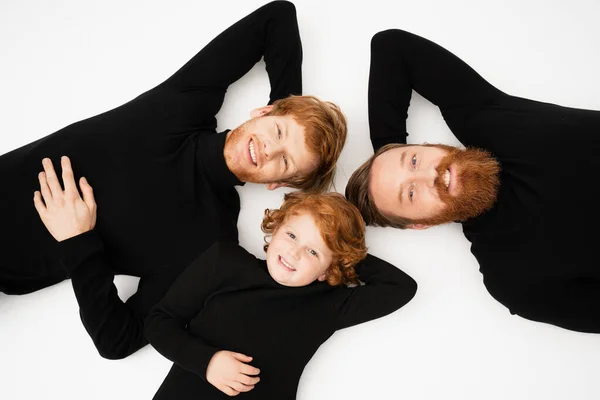 Top view of happy redhead men and boy in black sweaters looking at camera while lying on white background — Stock Photo