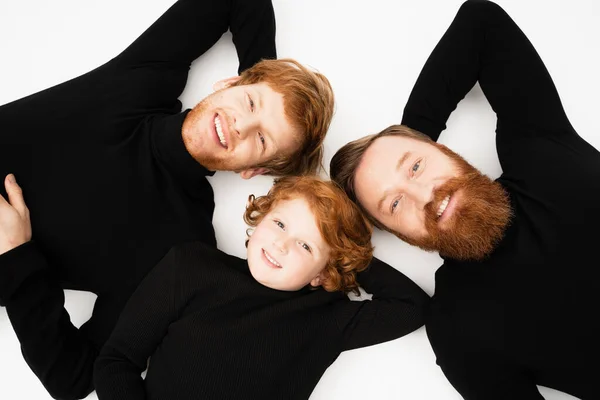 Top view of smiling redhead boy with bearded grandfather and dad in black turtlenecks lying on white background — Stock Photo