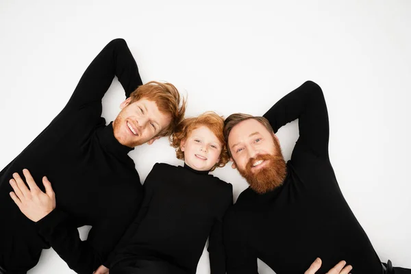 Top view of cheerful redhead boy with bearded granddad and father in black turtlenecks lying on light grey background — Stock Photo