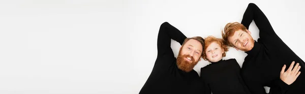 Top view of cheerful bearded man with red haired son and grandson smiling at camera while lying on light grey background, banner — Stock Photo