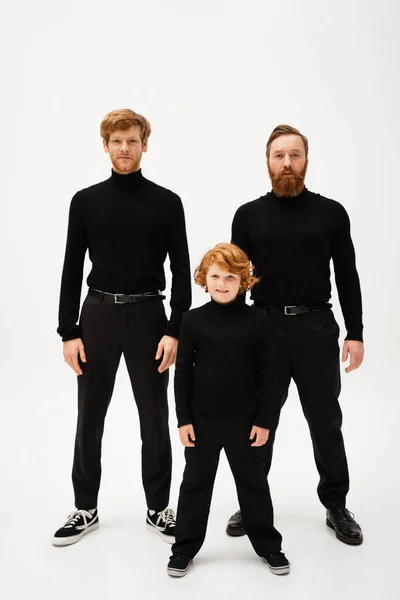 Full length of smiling boy and redhead bearded men in black turtlenecks and trousers standing on light grey background — Stock Photo