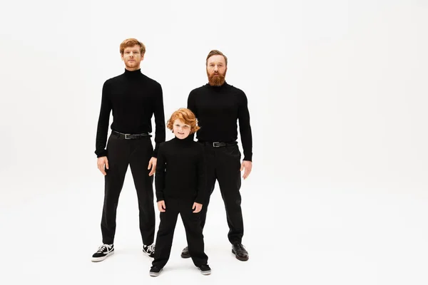 Full length of redhead men and kid in black sweaters and trousers standing and looking at camera on light grey background — Stock Photo