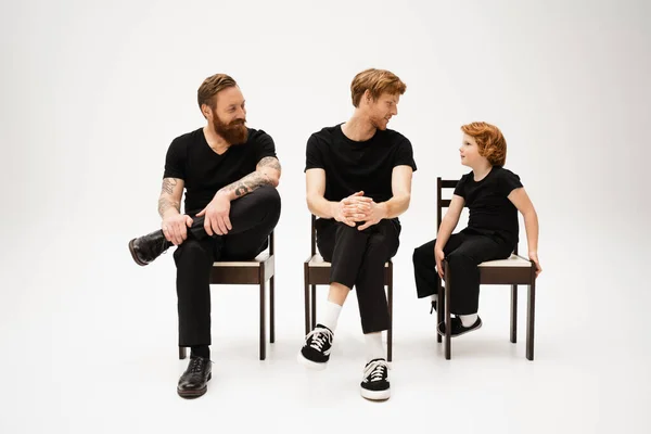 Redhead father and son looking at each other near smiling bearded man while sitting on chairs on grey background - foto de stock