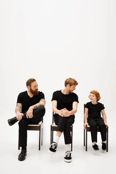 Bearded men in black clothes looking at smiling red haired boy while sitting on chairs on grey background — Stock Photo