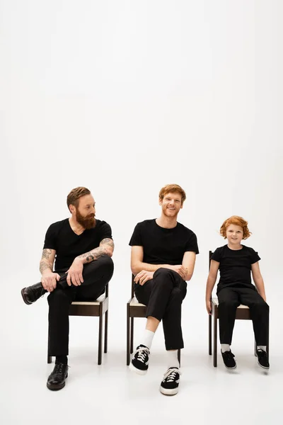 Cheerful redhead man smiling at camera near bearded tattooed dad and smiling son sitting on chairs on grey background — Stock Photo