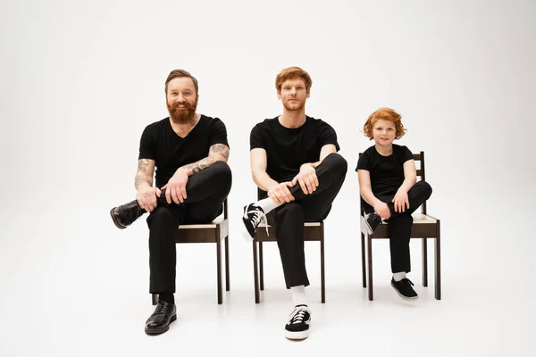 Joyful redhead boy with dad and tattooed grandpa sitting on chairs and smiling at camera on grey background — Stock Photo