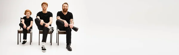 Full length of bearded men and redhead boy in black clothes sitting on chairs on grey background, banner — Stock Photo