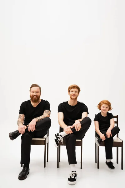 Cheerful red haired boy with bearded grandfather and dad sitting on chairs and smiling at camera on grey background — Stock Photo