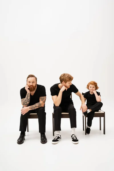 Bearded tattooed man smiling at camera near redhead son and grandson sitting on chairs on grey background — Stock Photo